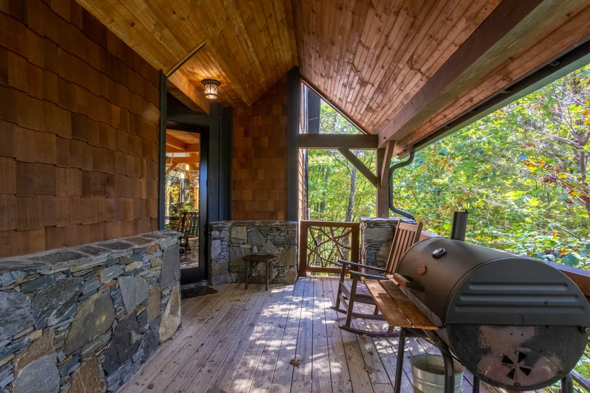 Featuring wooden rocking chairs on a porch, a stone wall, a grill, large windows, and lush surrounding greenery, this image embodies comfortable outdoor living. The scene conveys a sense of connection to nature and invites guests to relax, socialize, or simply enjoy the peace of their surroundings.