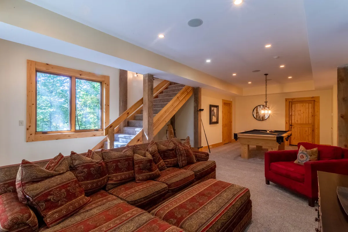 A comfortable lower-level room with a large sectional sofa facing a pool table. The space is designed for both socializing and individual relaxation, with plenty of room for families or groups to spread out. Warm lighting and rustic elements contribute to an atmosphere perfect for game nights or quiet reading.