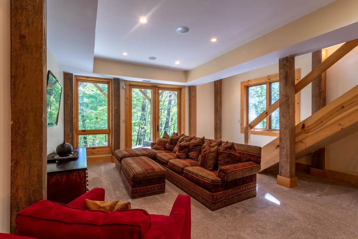 A spacious living room featuring a large sectional couch positioned by floor-to-ceiling windows. The windows provide abundant natural light and offer glimpses of the outdoors, while a wooden staircase adds rustic charm. The inviting seating area suggests a comfortable space for relaxing after outdoor adventures.