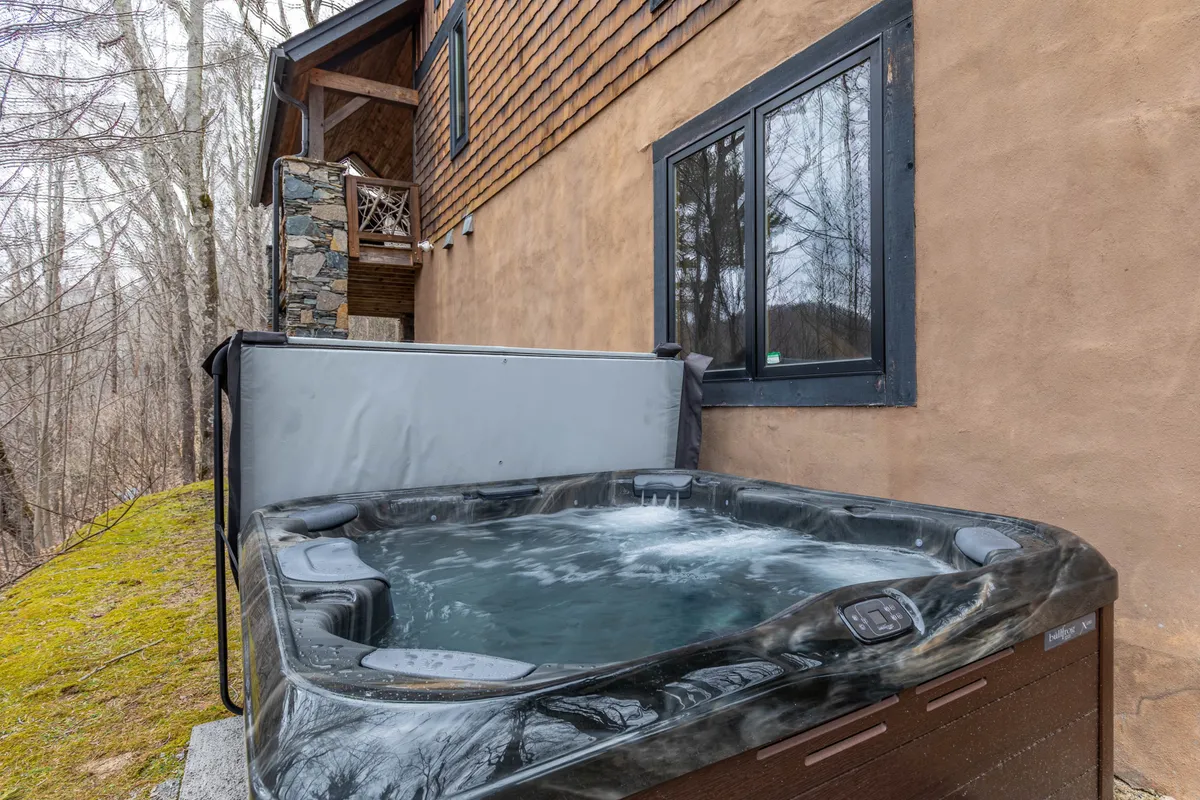 An inviting outdoor hot tub sits next to a stone wall and wooden balcony, nestled in nature. The setting offers privacy and scenic views, making it ideal for unwinding under the stars or enjoying the fresh mountain air after indoor fun.