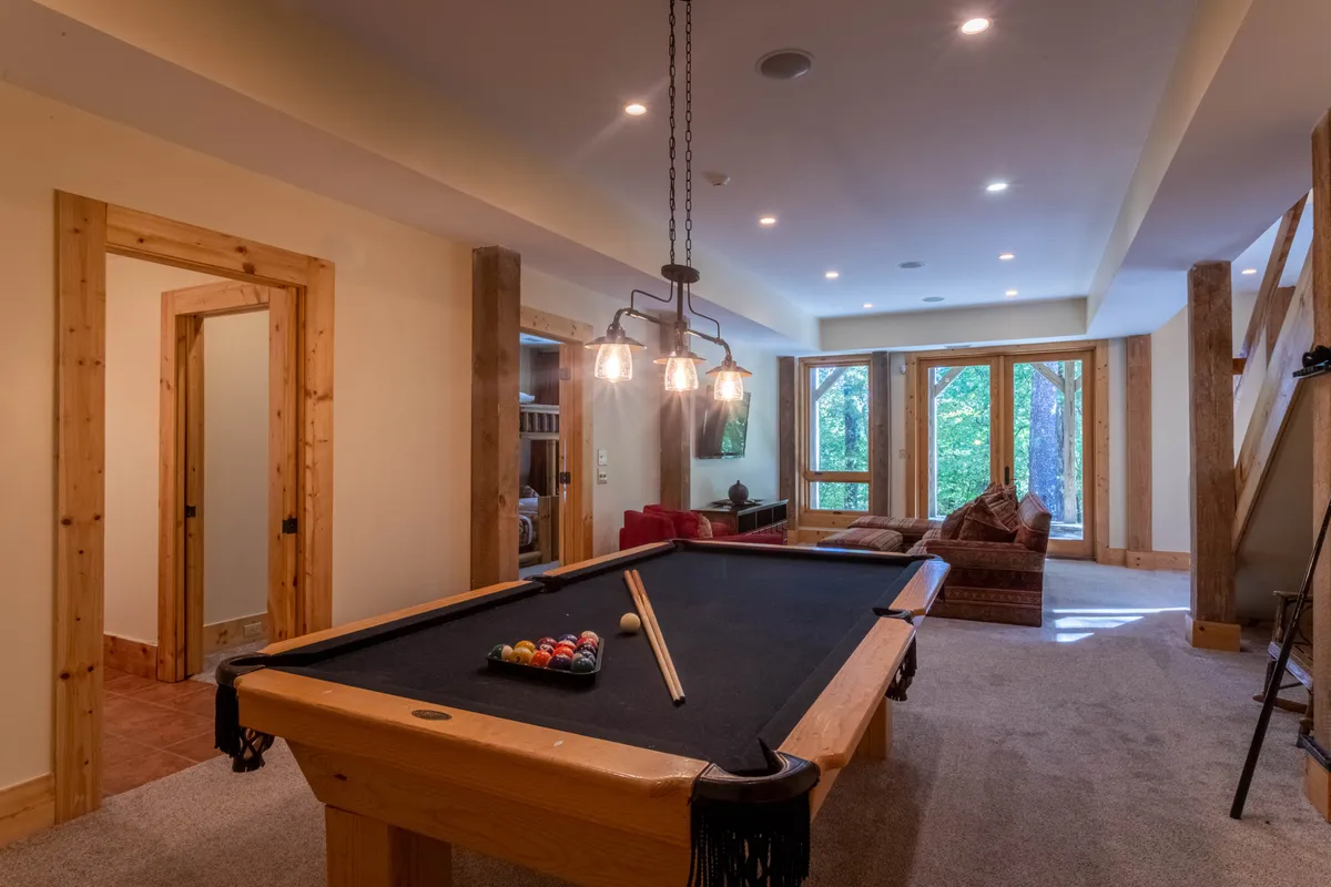 A spacious room featuring a pool table set near large windows that flood the room with natural light. The setting includes comfortable sofa seating and exposed wooden beams, creating a social and relaxed atmosphere with views of the outdoors visible through the windows.