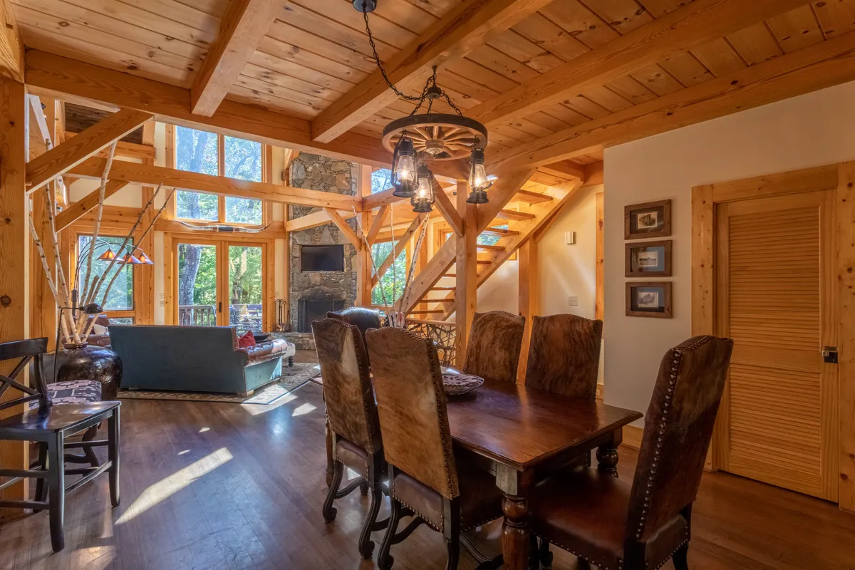 A spacious interior scene encompassing a dining table, visible wooden beams, a fireplace, and large windows. The layout suggests openness, with clear sightlines connecting dining and living spaces. Family and friends are shown gathered for meals and relaxation, reinforcing the theme of shared experiences.