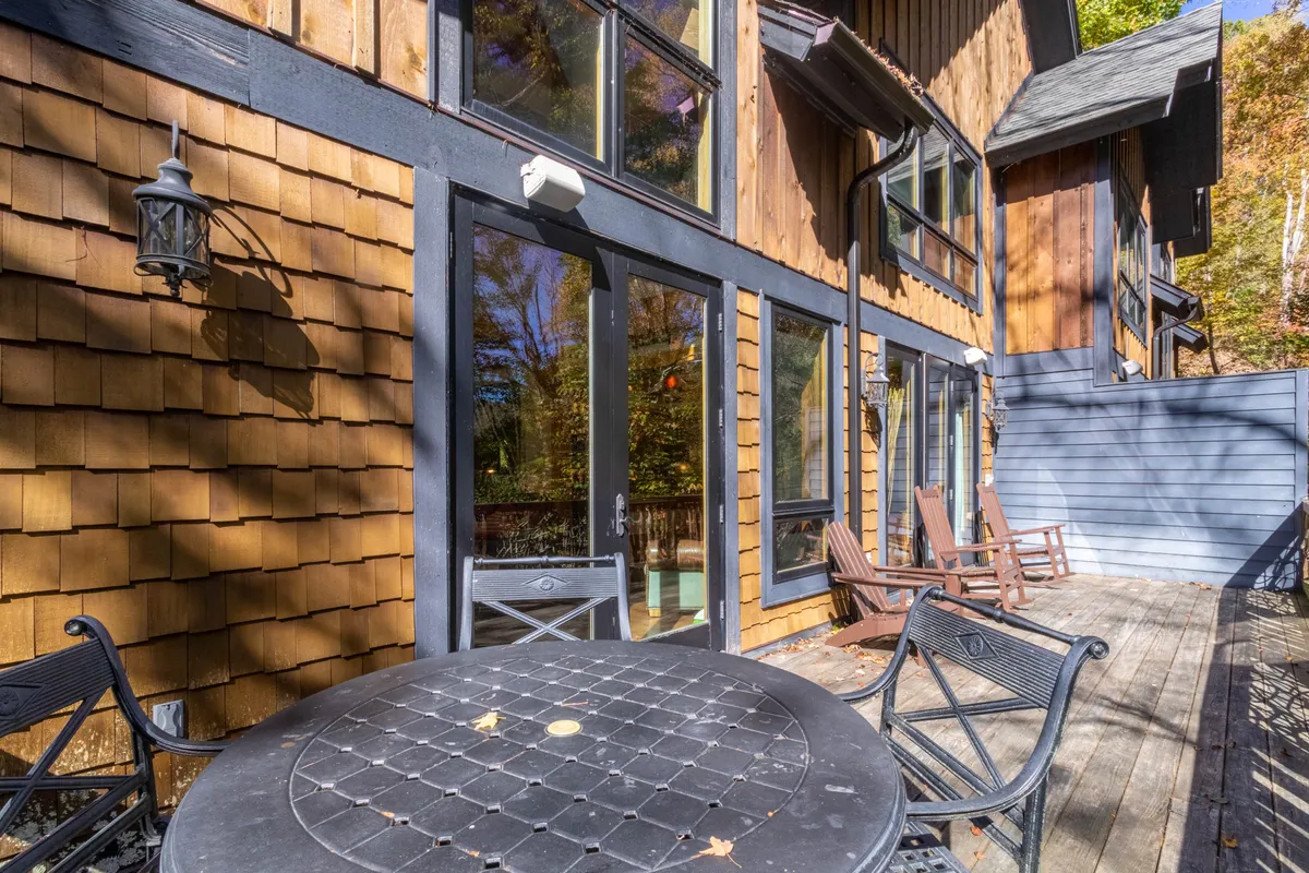 This image showcases a welcoming wooden deck outfitted with a round table, rocking chairs, and large windows looking into the home. It offers a seamless transition between indoor luxury and the natural mountain setting, highlighting opportunities for outdoor dining, relaxation, and socializing with picturesque views.
