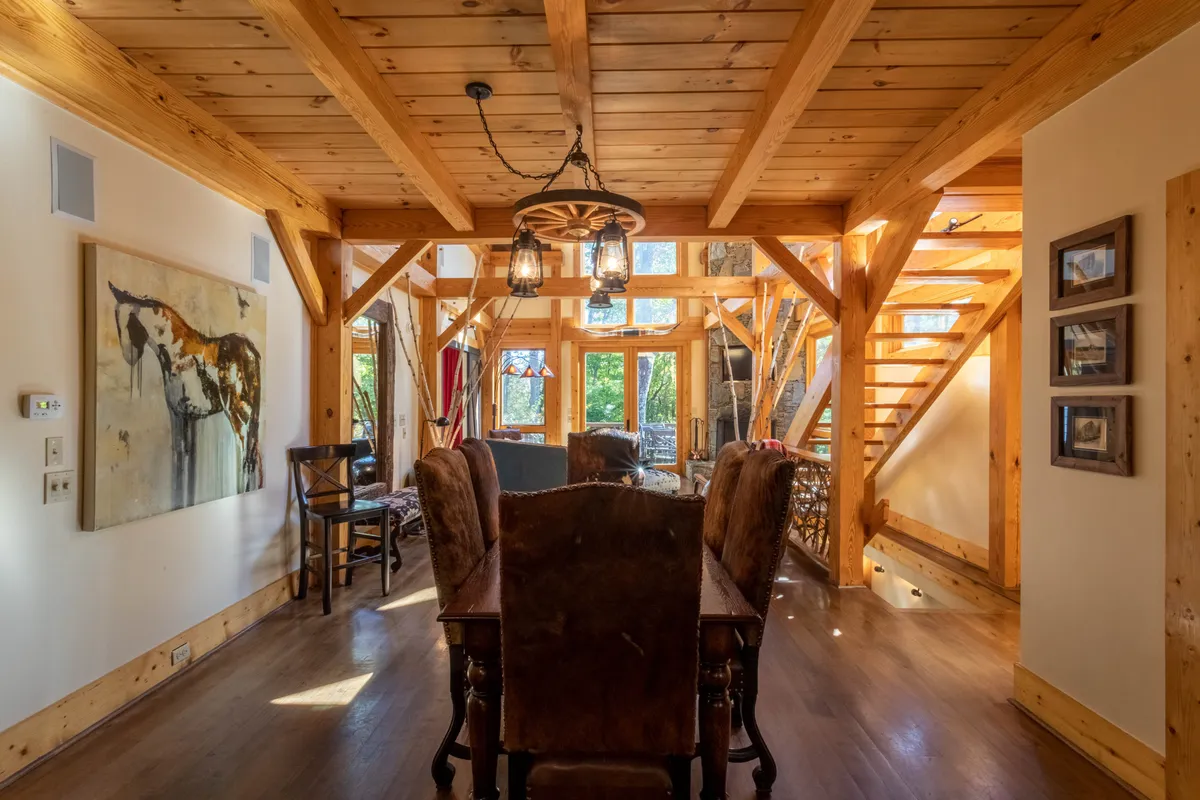 A sunlit dining space with exposed wooden beams, a dining table ready for gatherings, unique artwork on the wall, and a staircase leading to other areas. The atmosphere is warm and inviting, ideal for sharing meals and stories.