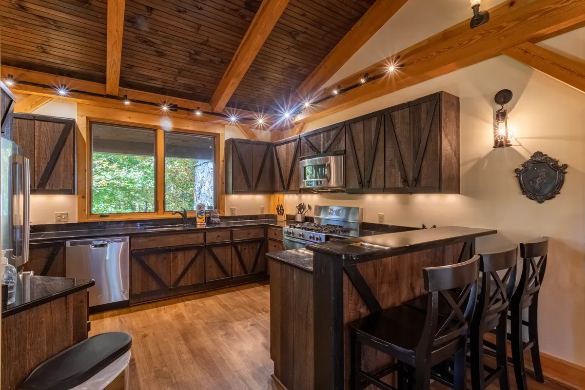 A bright, welcoming kitchen highlighted by a large kitchen island, wooden beams, a prominent large window, and stainless steel appliances. The atmosphere is rustic yet modern, with ample space for gathering, cooking, and entertaining.