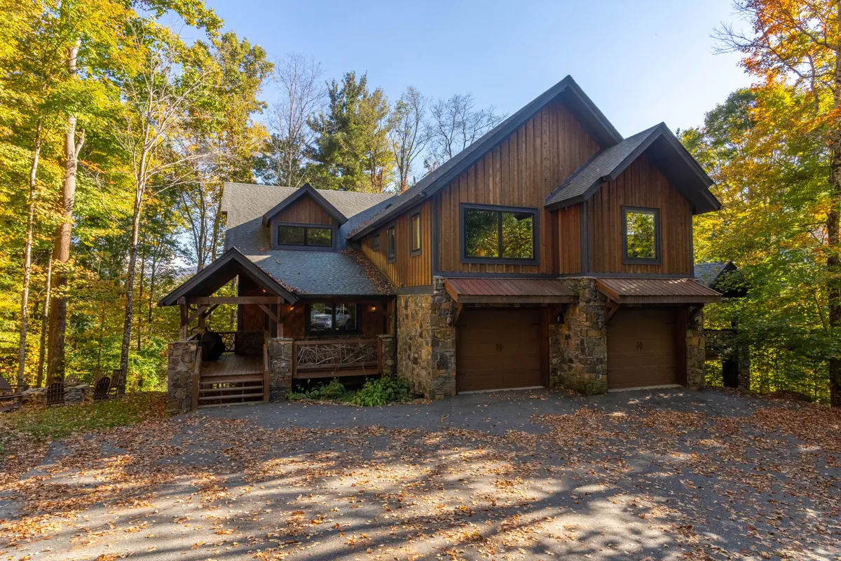 The image showcases the inviting wooden and stone exterior of the Rustic Cedar Rock Townhome, nestled among towering trees. Expansive windows promise breathtaking mountain vistas, while a spacious porch invites guests to unwind and enjoy the sunset, all set against a backdrop of lush nature.