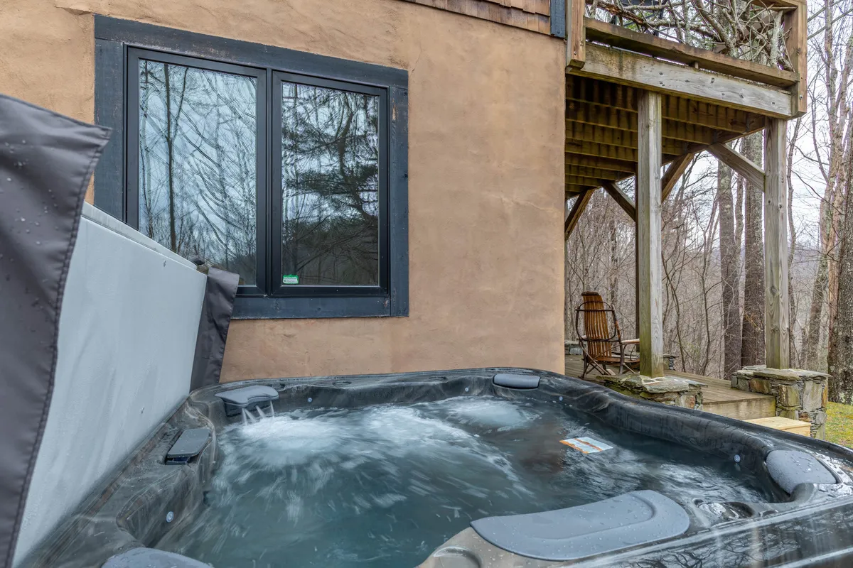 A serene hot tub on a wooden deck, surrounded by tall trees, with a nearby window hinting at the cozy home. The setting is ideal for unwinding after action-packed days, with nature close at hand.